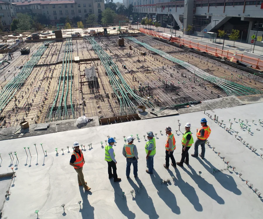 A construction crew in hi-vis vests and hard hats reviewing progress on an active building site, seen from above.