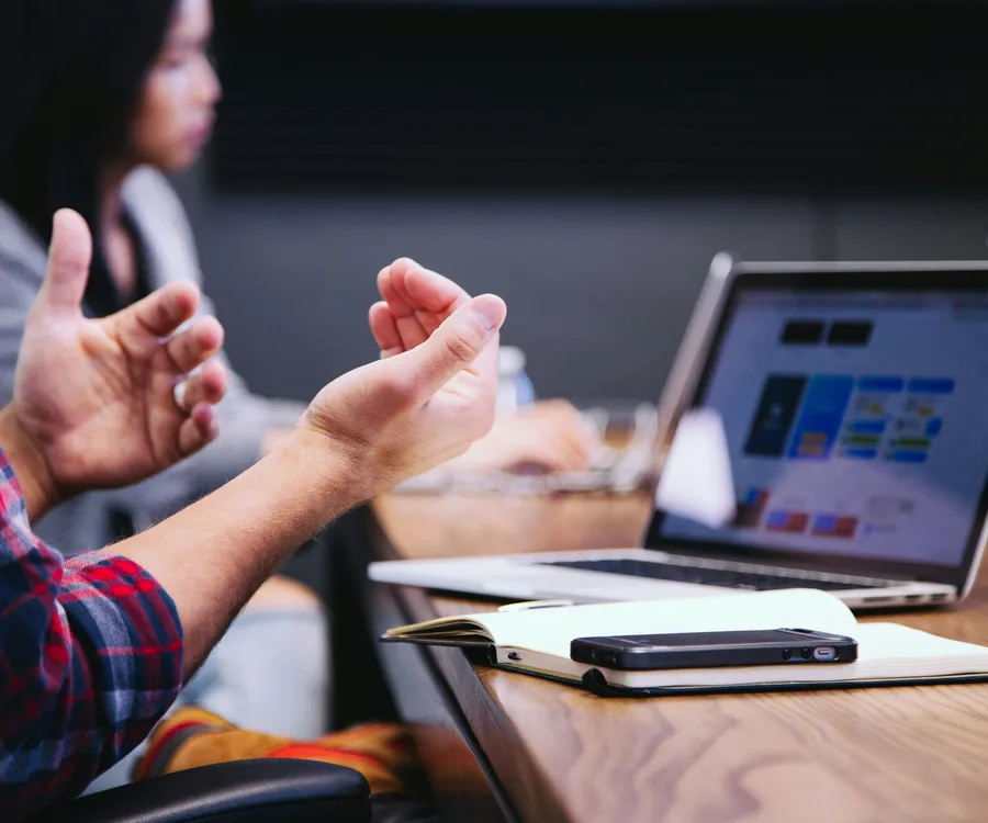 A consultant gesturing mid-discussion in a meeting, laptop open on the table showing a project dashboard.