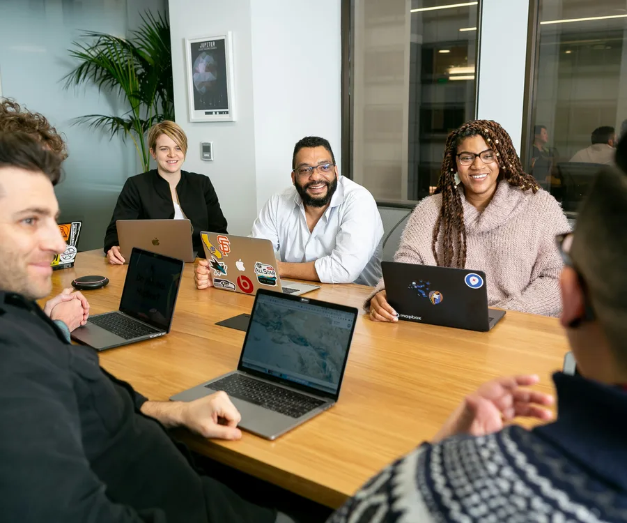 An engineering team gathered around a conference table, laptops open, mid-discussion in a standup-style meeting.
